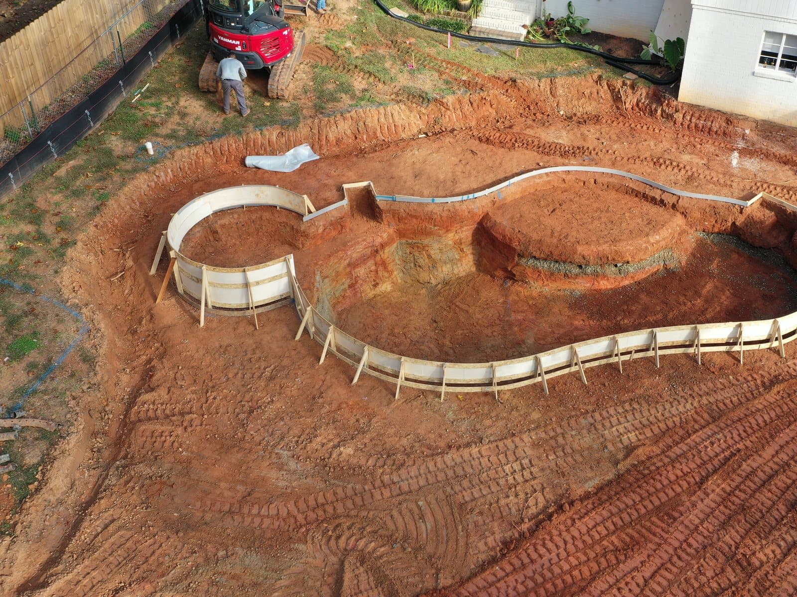 A pool shell formed in red clay, seen from above, the excavator still working at the house