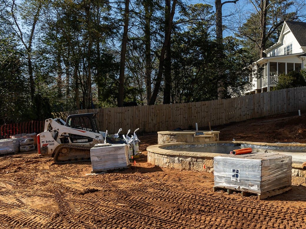 A shrink-wrapped pallet of stone coping staged at the edge of a finished pool shell, the raised spa behind it and the excavator still working at the house